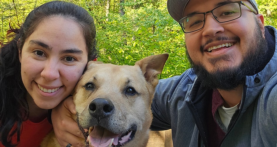 A photo of two people smiling outdoors with Scooter, a Carolina dog crossbreed.